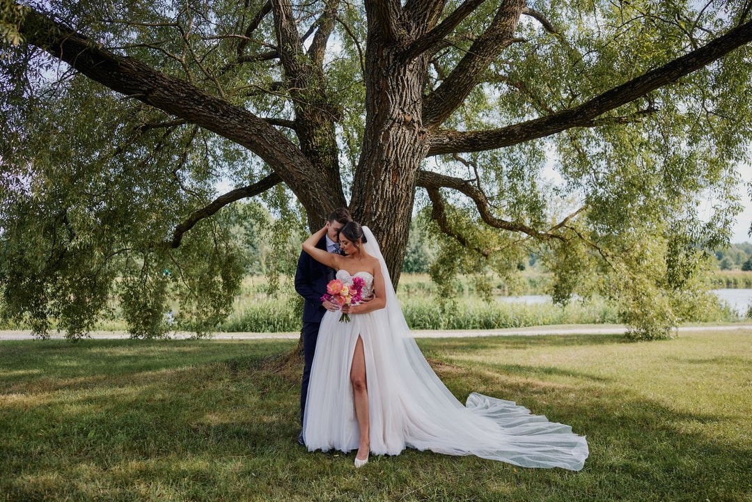 Para młoda pod starym drzewem, panna młoda w białej sukni z trenią trzyma bukiet ślubny w jasnej palecie, jezioro w tle