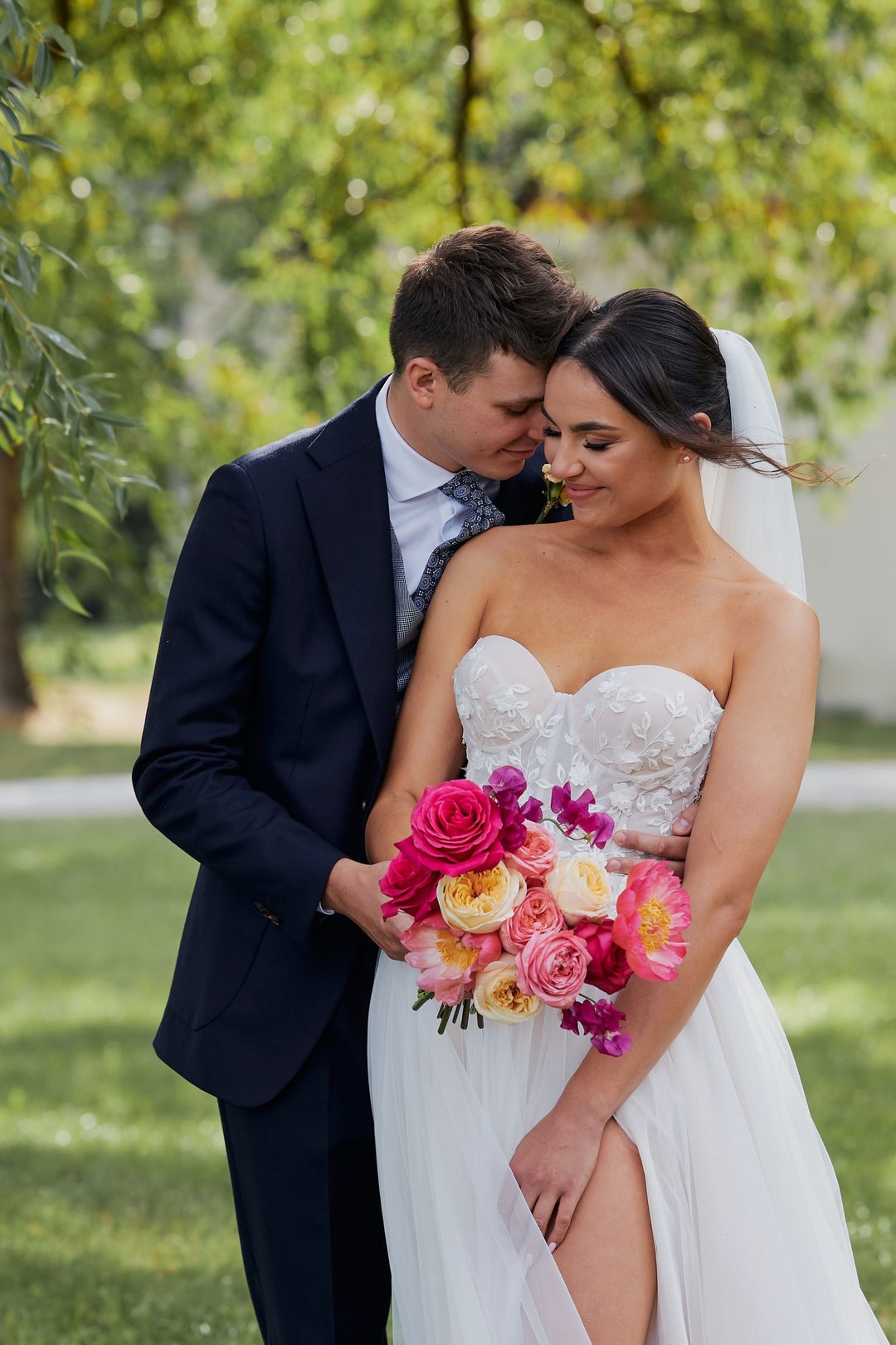 Para Młoda w ogrodzie, mężczyzna w ciemnym garniturze całuje kobietę w białej sukni ślubnej, która trzyma bukiet ślubny z różowymi i żółtymi różami.