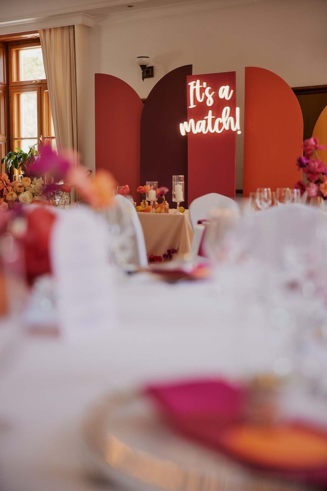 Dekoracja sali weselnej - czerwone serca z napisem "It's a match" na tle, stół z białym obrusem, kompozycje kwiatowe w ciepłych tonach.