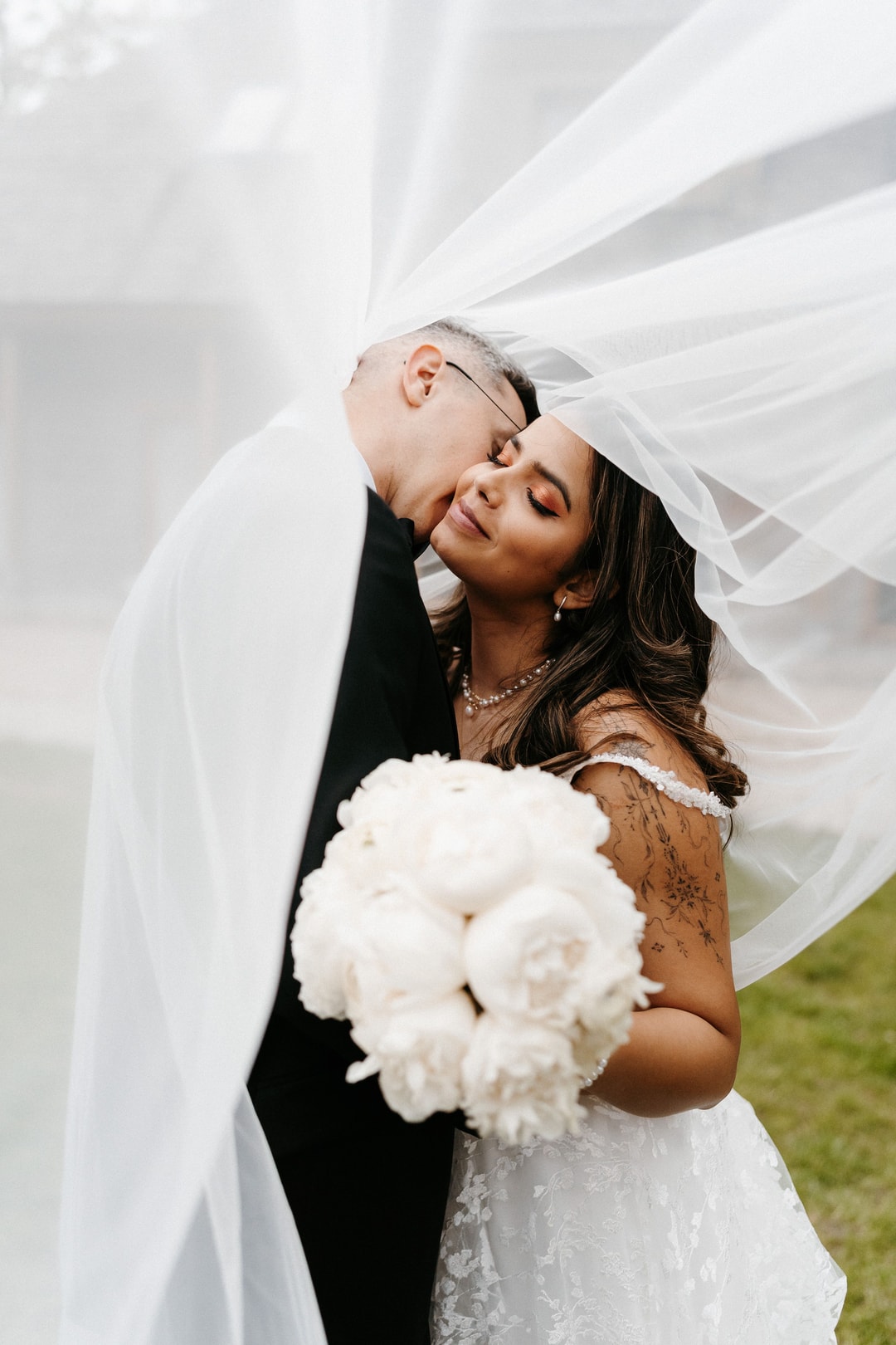 Para Młoda z bukietem ślubnym - mężczyzna w czarnym garniturze całuje kobietę w białej sukni z welona, kobieta trzyma białe róże