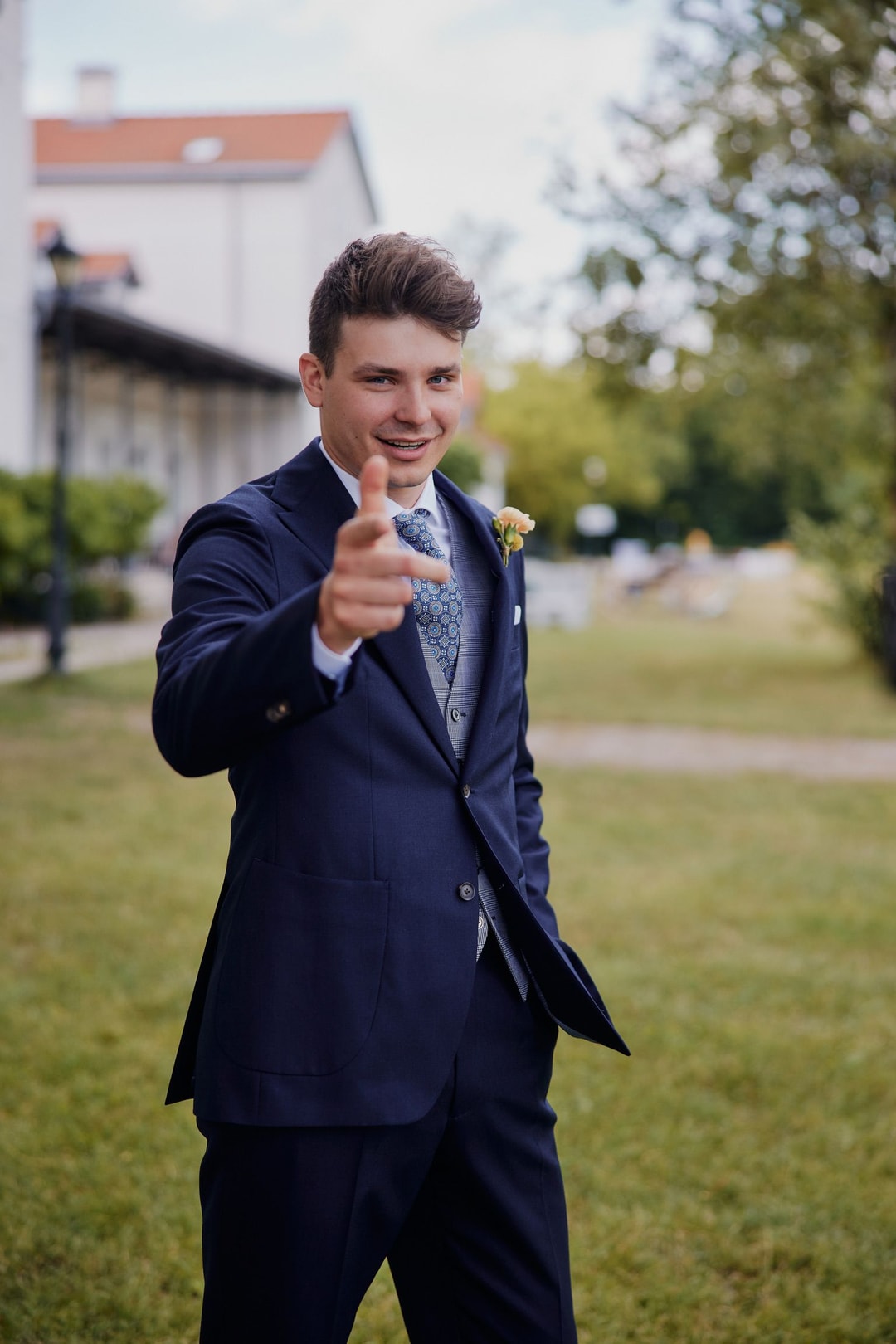 Młody mężczyzna w granatowym garniturze z butonierką, wskazujący palcem w kamerę, na tle zieleni i budynku.