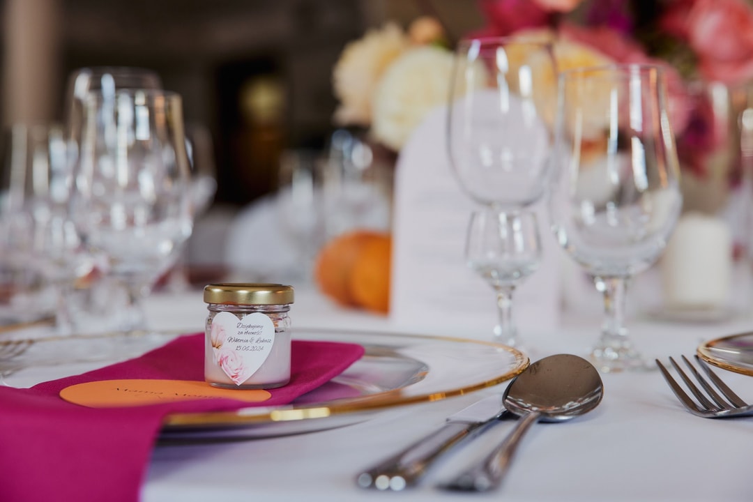 Dekoracja stołu weselnego z białym talerzykiem, złotą obwódką, różowym serwetką, szklankami i małym słoiczkiem z różową etykietą