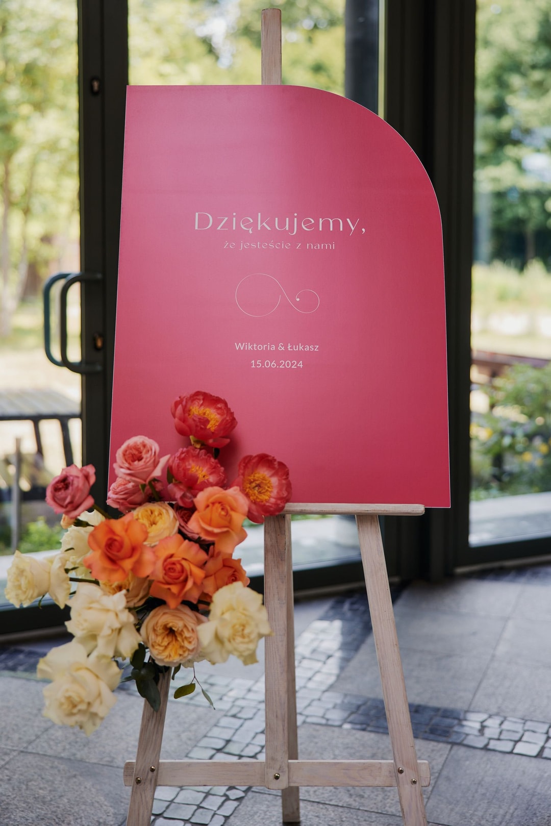 Tablica welcome z napisem "Dziękujemy, ze jesteście z nami" i datą, dekorowana kwiatami w ciepłych tonach przy wejściu.