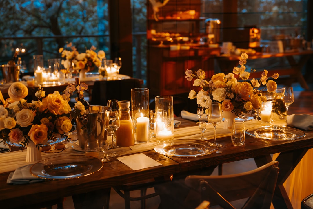 Drewniany stół z dekoracją stołu – złote i pomarańczowe kwiaty w wazonach, świece, szklanki i nakrycia