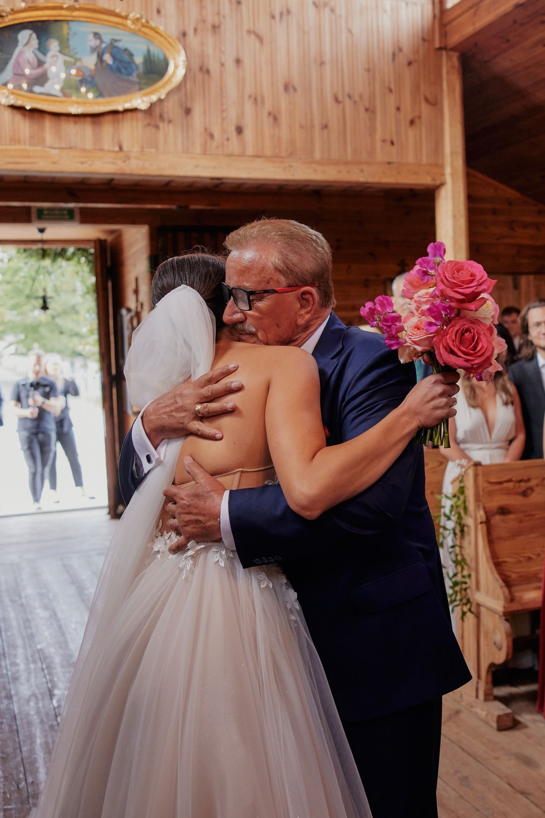 Mężczyzna w garniturze przytula pannę młodą w białej sukni, trzymając bukiet ślubny z różami w odcieniach różu i czerwieni, drewniana sala weselna