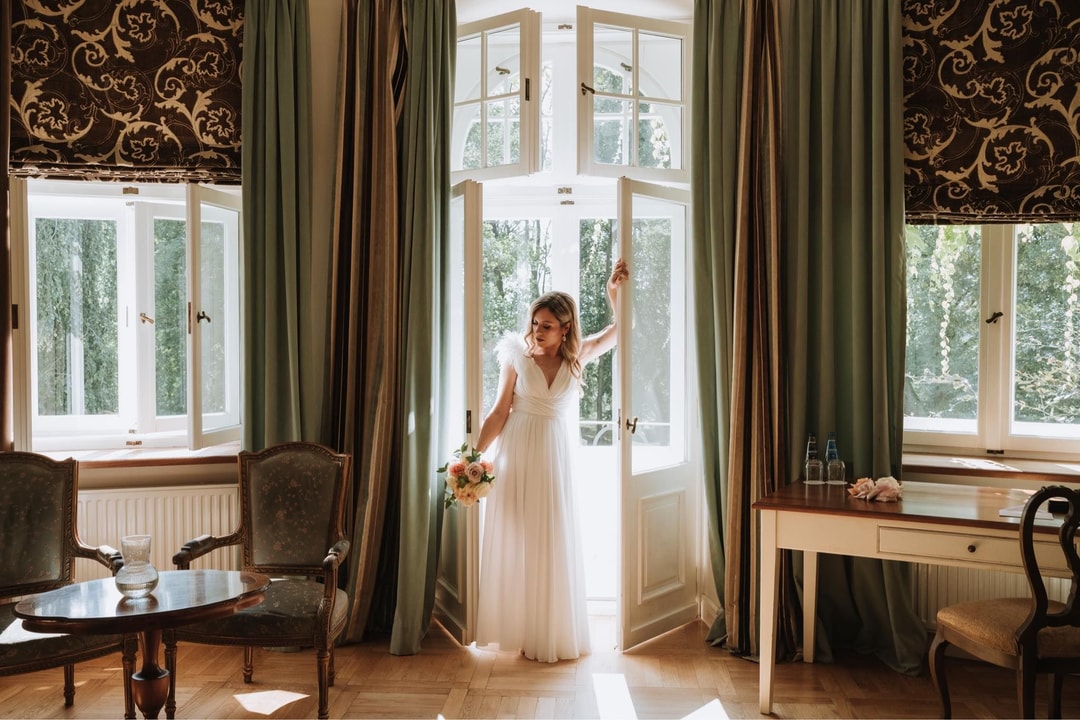 Panna młoda w białej sukni trzymająca bukiet ślubny, stojąca przy otwartych drzwiach balkonowych, elegancka sala z zieloną tkaniną i ornamentacyjnym panelem.