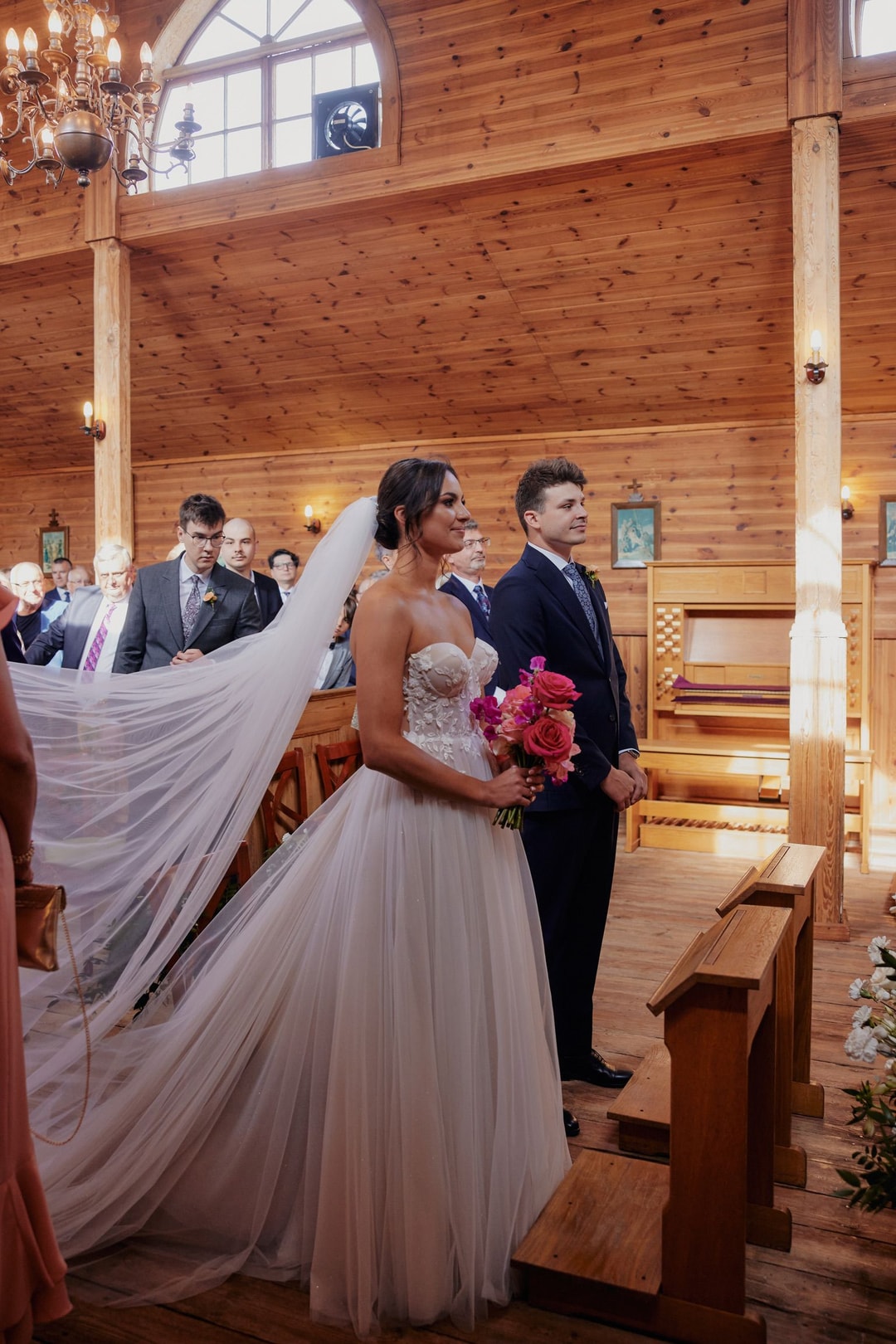 Para Młoda podczas ceremonii w drewnianej kaplicy, bukiet ślubny z różowymi kwiatami, goście w tle