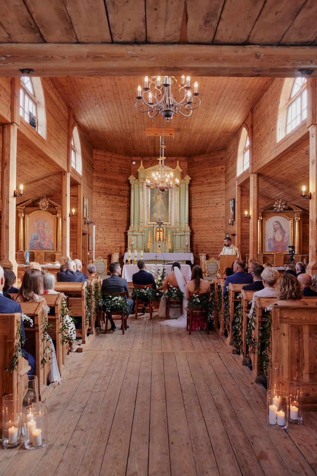 Drewniana kaplica z dekoracjami sali weselnej: zielone girlandy przy ławkach, świece, elegancki żyrandol, ołtarz ze złotymi zdobieniami, goście siedzą w ławkach.