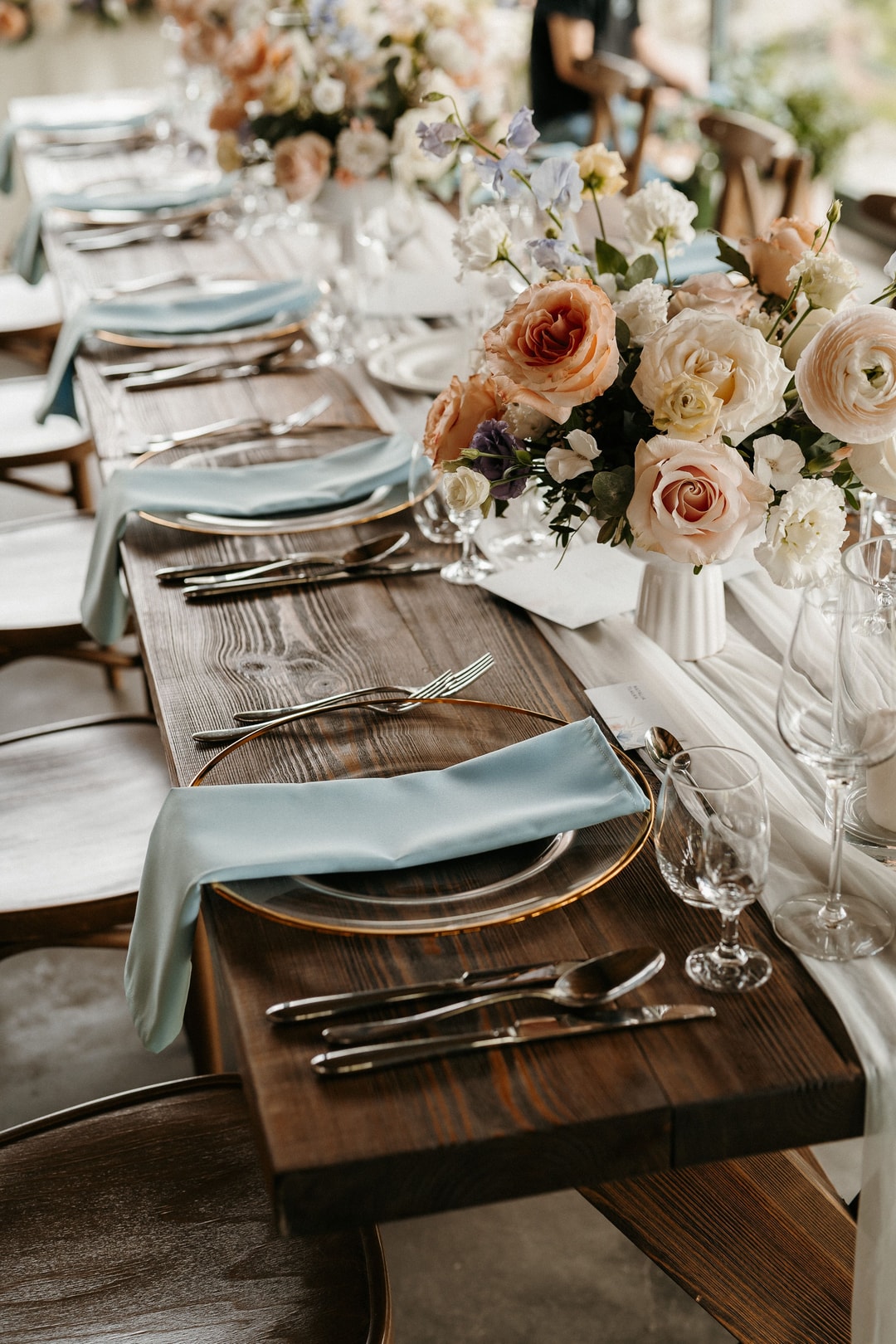 Dekoracja stołu weselnego z kompozycją kwiatową w wazie, talerze, sztućcach i serwecie w pastelowych tonach