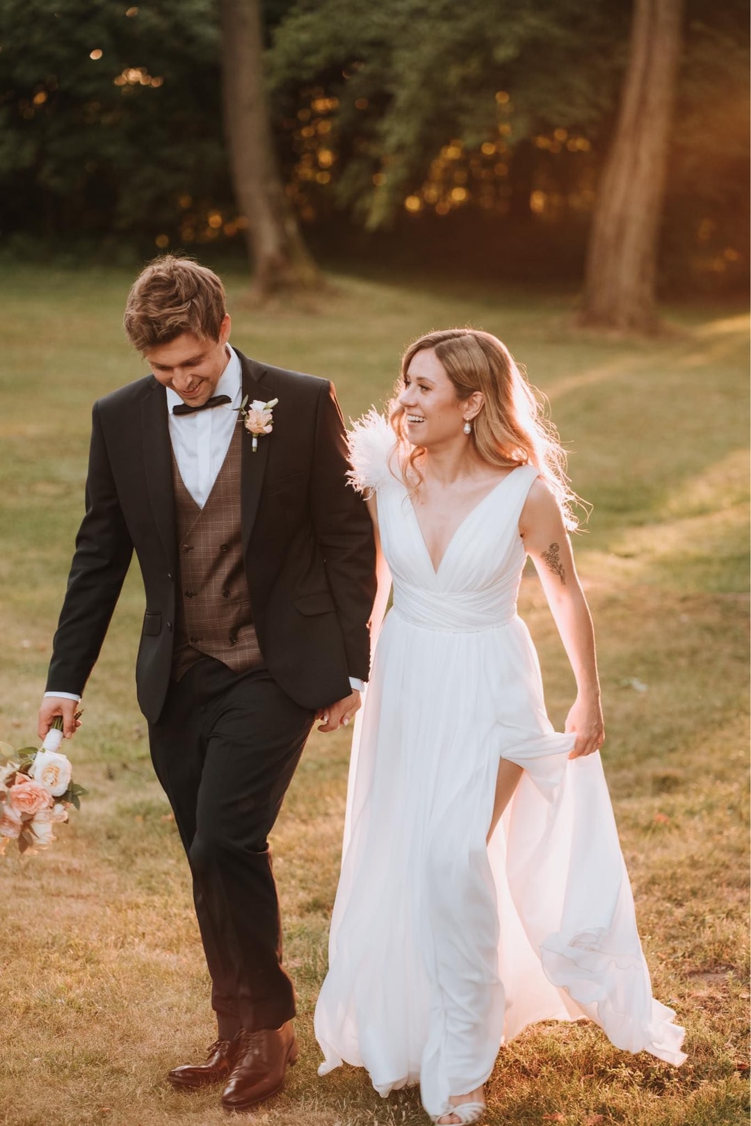 Para Młoda spaceruje po trawniku, mężczyzna w czarnym garniturze trzyma bukiet ślubny, kobieta w białej sukni ślubnej, drzewa w tle.