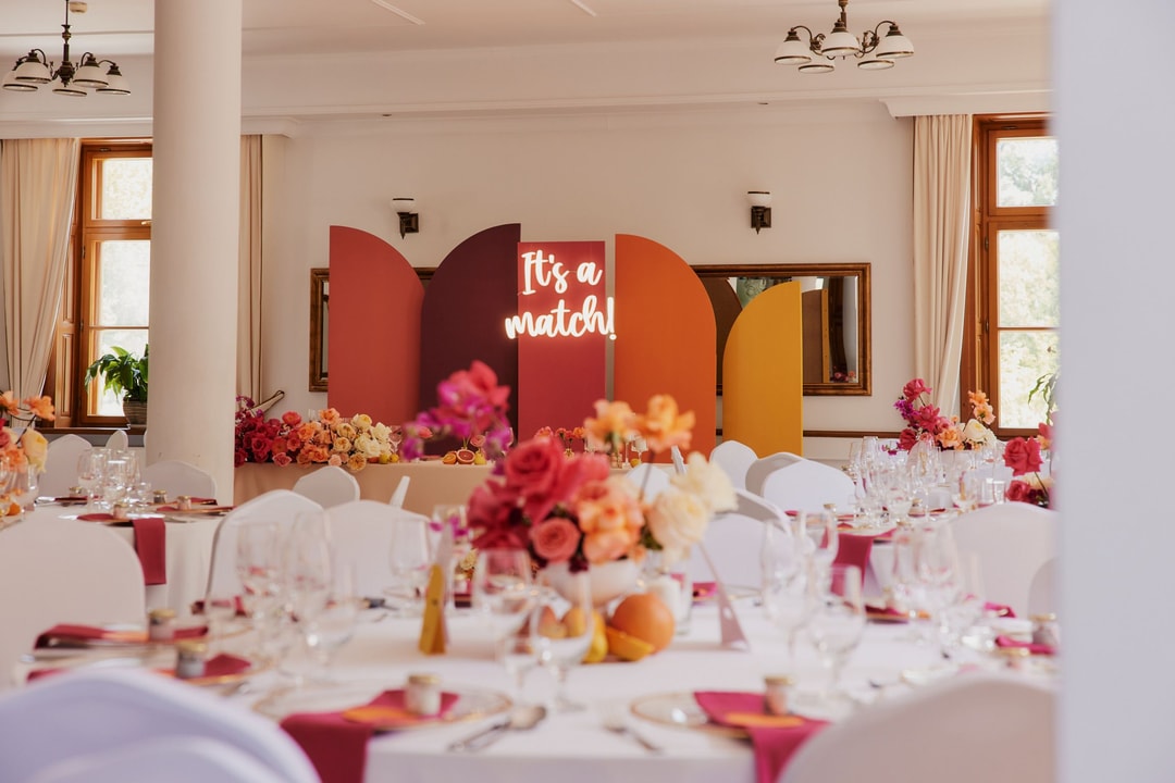 Aranżacja stołów weselnych z białymi krzesłami, kolorowymi kwiatami w różu, pomarańczu i czerwieni, oraz dekoracyjnym panelem z napisem "It's a match" w tle