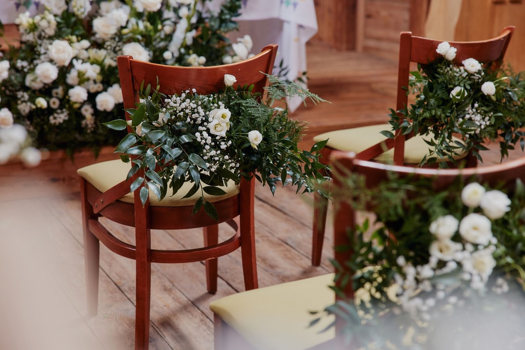 Drewniane krzesła z białymi poduszkami i kompozycjami kwiatowymi na aranżację stołów, białe kwiaty z zielenią w tle