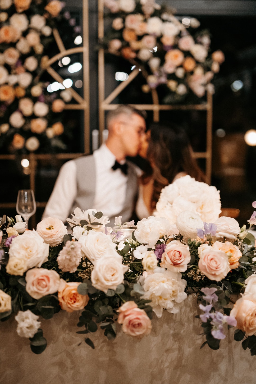 Kompozycja kwiatowa na stół z białymi i łososiowymi różami, piwoniami i zielenią. W tle para w strojach ślubnych przy dekoracji sali weselnej.