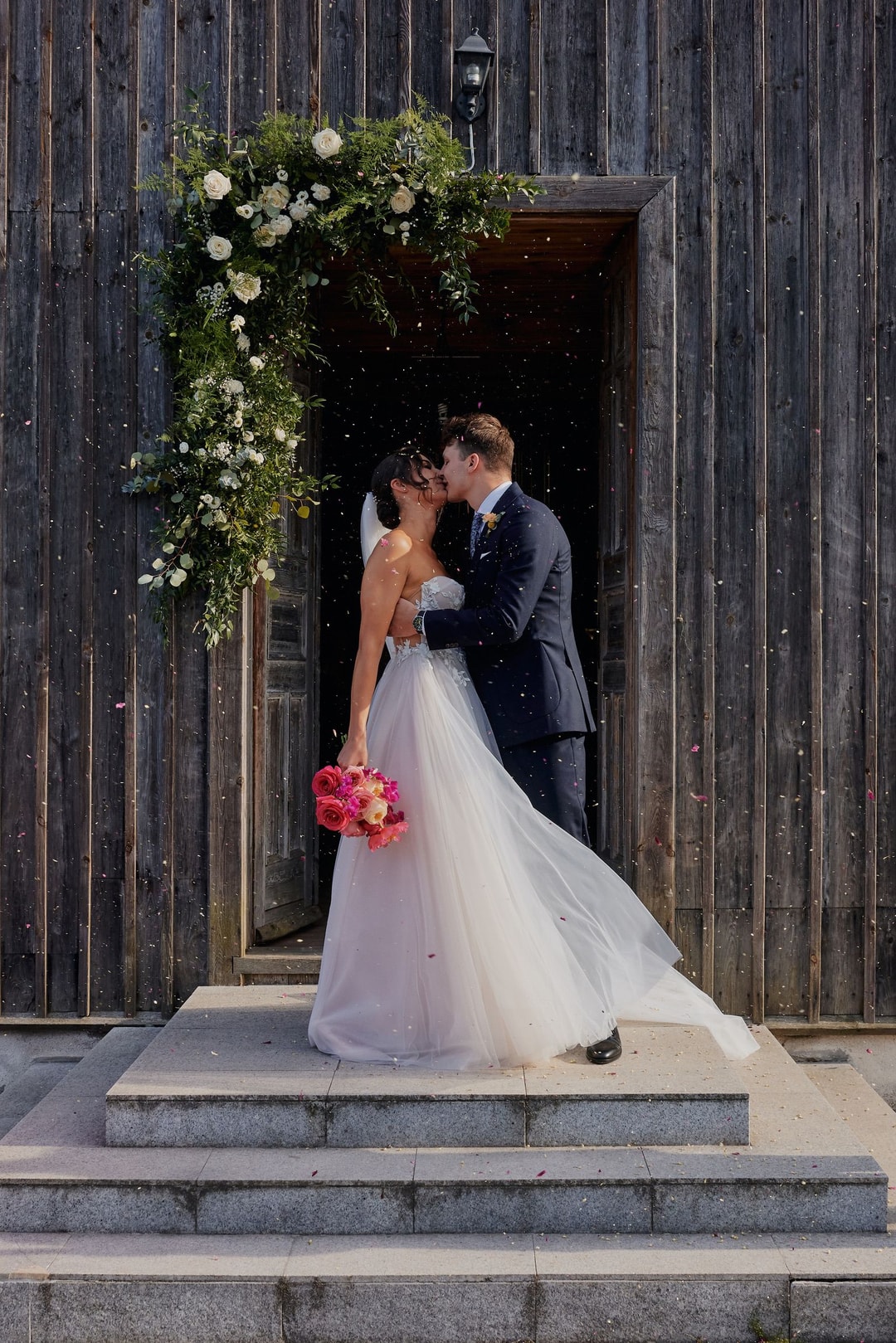 Para Młoda całuje się w wejściu z łukiem kwiatowym. Dekoracja z białych kwiatów i zieleni przy drewnianym wejściu, pada konfetti.