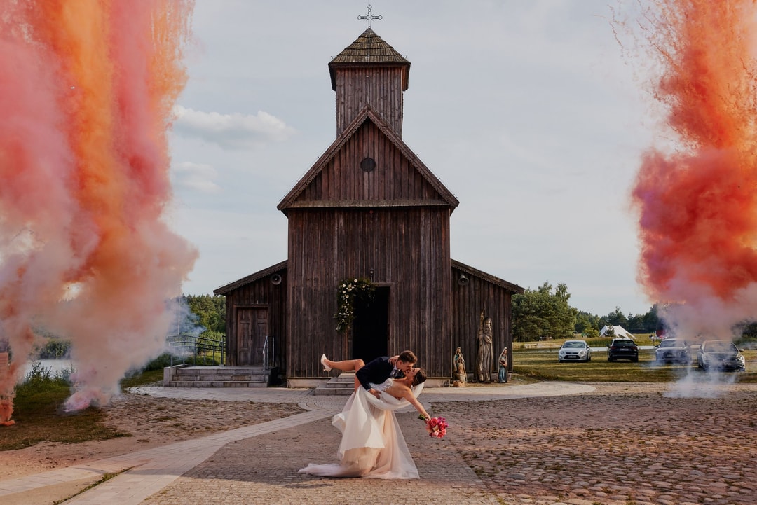 Kobieta w białej sukni trzymająca bukiet czerwonych kwiatów przed drewnianą kaplicą z kolorowymi dymami po obu stronach