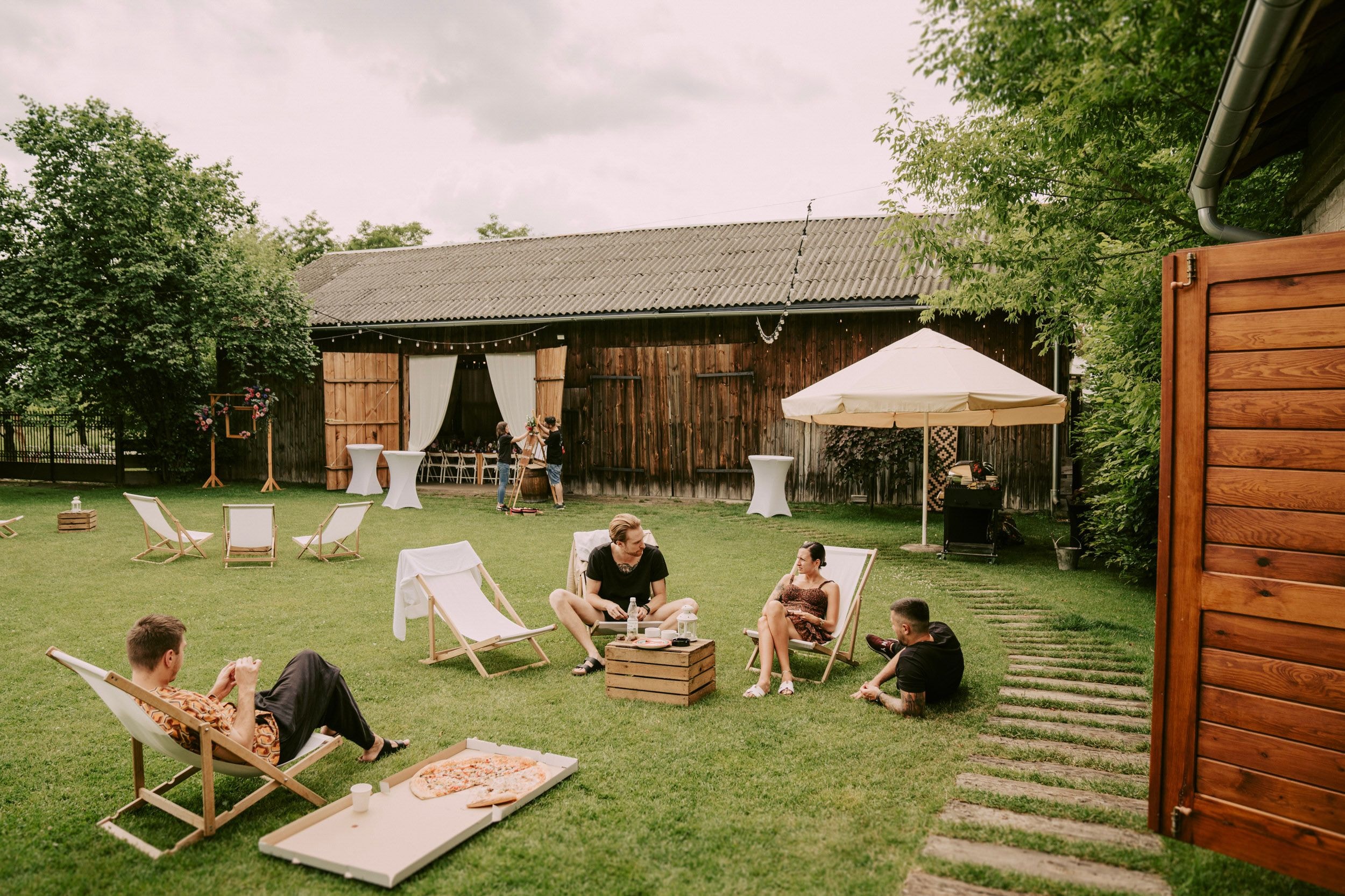 Grupa ludzi relaksuje się na podwórku z zieloną trawą i drzewami. Są tam leżaki i stół z napojami. W tle widać drewniany budynek i parasol ogrodowy, tworząc przytulne otoczenie na zewnątrz.