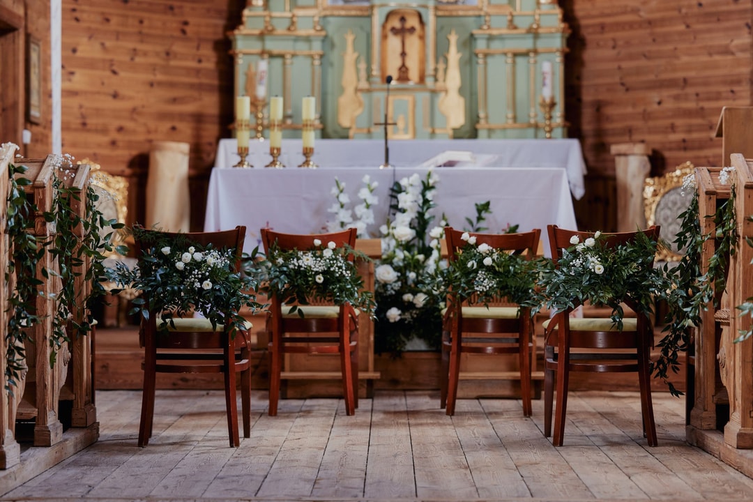 Drewniana sala z drewnianymi krzesłami ozdобionymi girlandami zieleni i białych kwiatów, aranżacja stołów weselnych z ołtarzem w tle