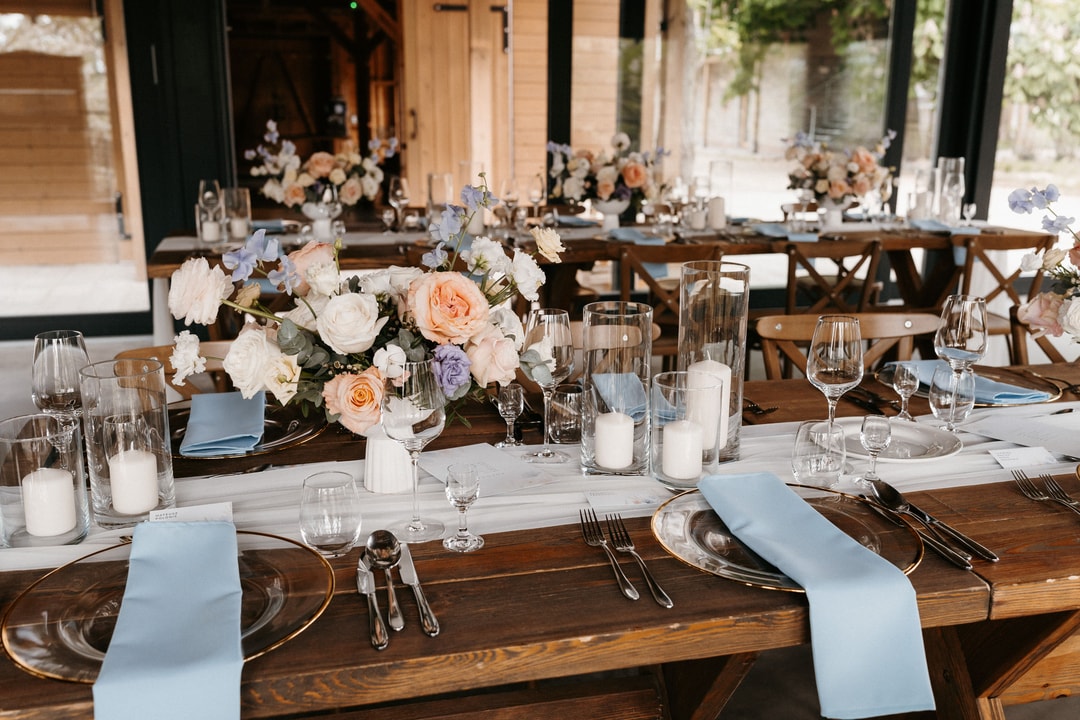 Aranżacja stołów weselnych z drewnianymi blatami, dekoracja kwiatowa w pastelowych tonach, błękitne serwetki, świece i kieliszki