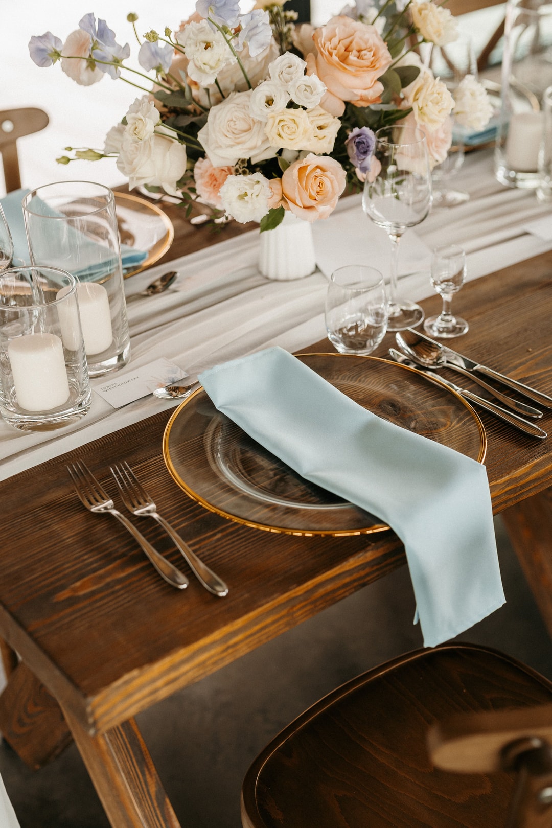 Dekoracja stołu z kompozycją kwiatową w białej wazie, pełną róż i delikatnych kwiatów w jasnej palecie, obok ustawione kieliszki i sztućce na drewnianym blacie.