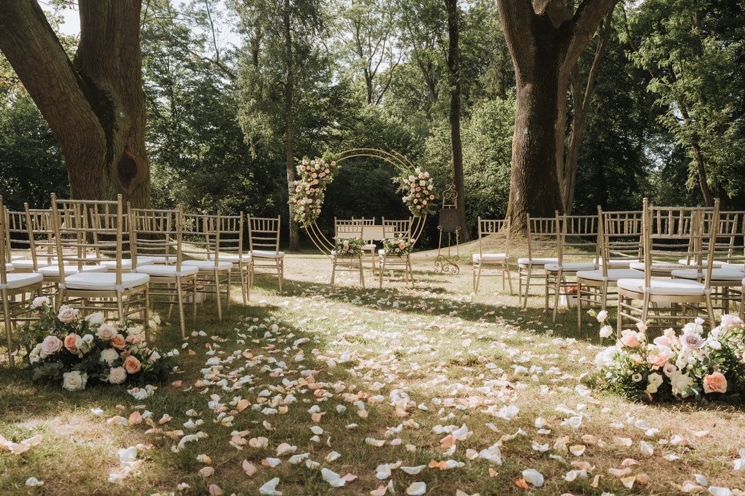 Łuk kwiatowy z różami w pastelowych tonach, drewniane krzesła gościnne, dekoracje sali weselnej z płatkami kwiatów na ziemi, drzewa w tle