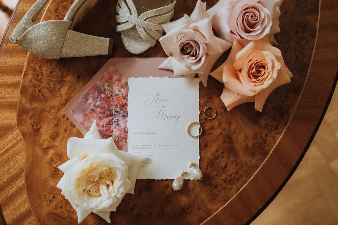 Drewniany stół z kompozycją kwiatową na stół: róże w pastelowych tonach, papeteria ślubna, obrączki i złote buty ślubne