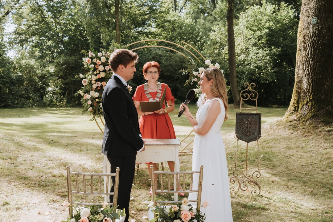 Łuk kwiatowy z różami i białymi kwiatami, ceremonia ślubna w ogrodzie z parą młodą i celebrantką.