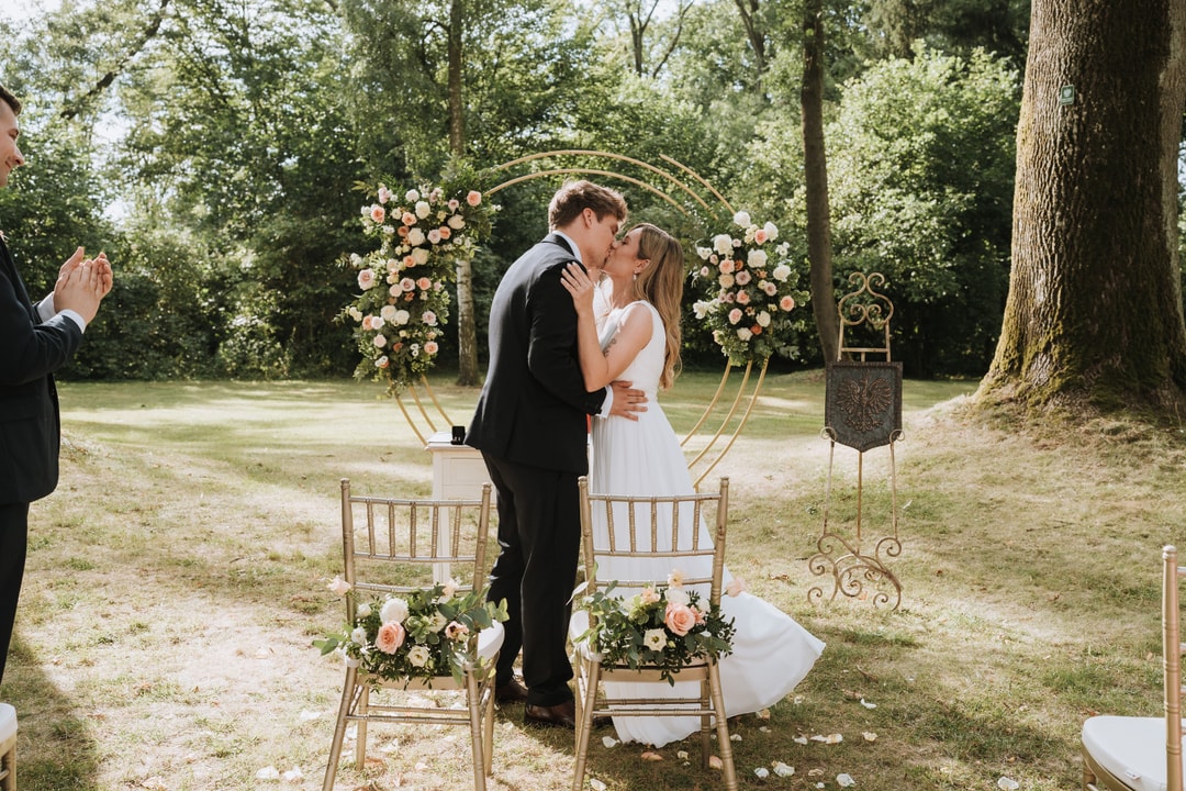 Para Młoda całująca się pod łukiem kwiatowym z różami i białymi kwiatami w ogrodzie, goście klaskują.