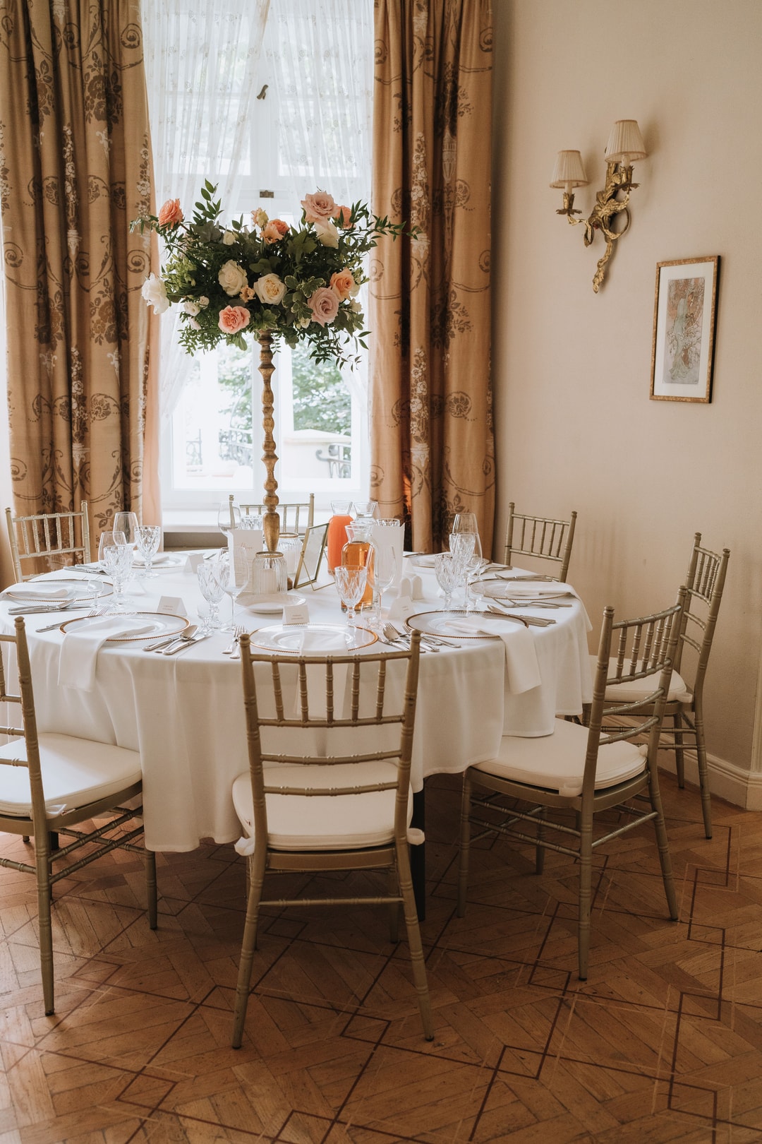 Dekoracja stołu weselnego z białym obrusem, композycją kwiatową na postumencie z różami w jasnej palecie, złotymi naczyniami i szklankami