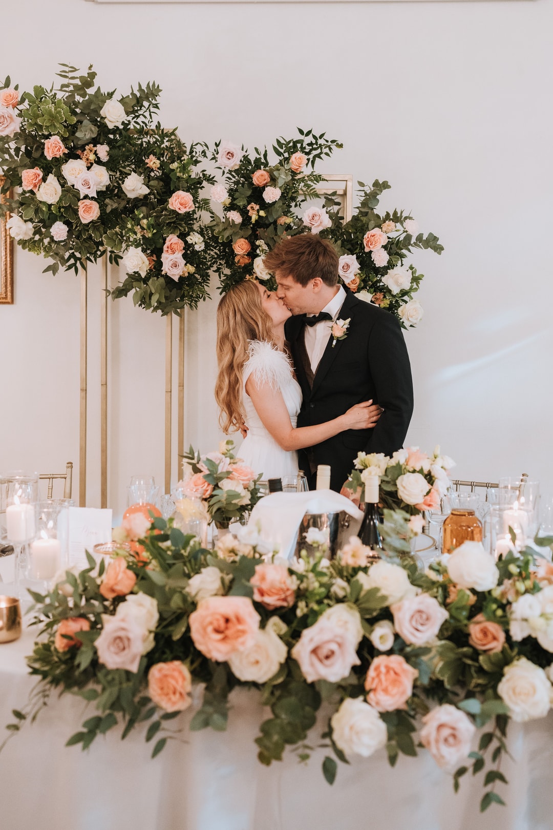 Para w strojach ślubnych całuje się przy łuku kwiatowym z różami w jasnej palecie i zielenią, dekoracja sali weselnej.