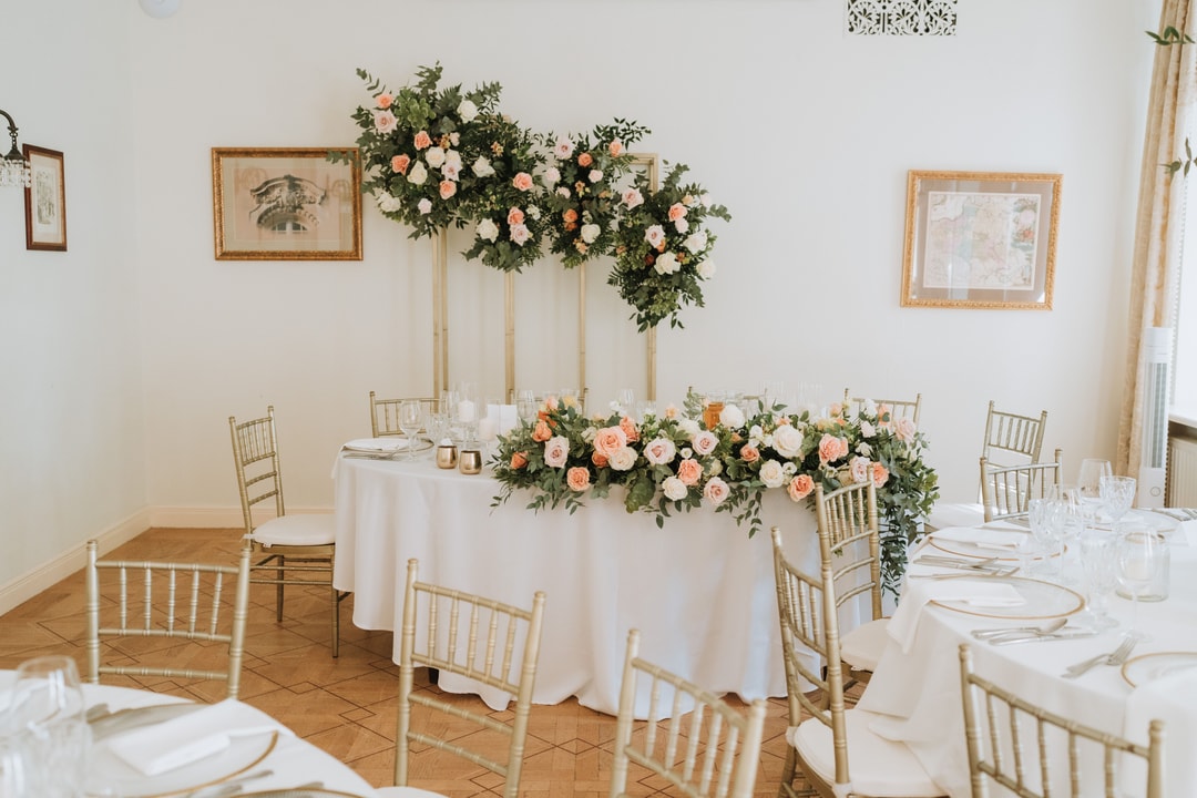 Dekoracje sali weselnej z okrągłym stołem, białym obrusem, złotymi naczyniami i kompozycją kwiatową z kwiatów w jasnej palecie, otoczoną zielenią.