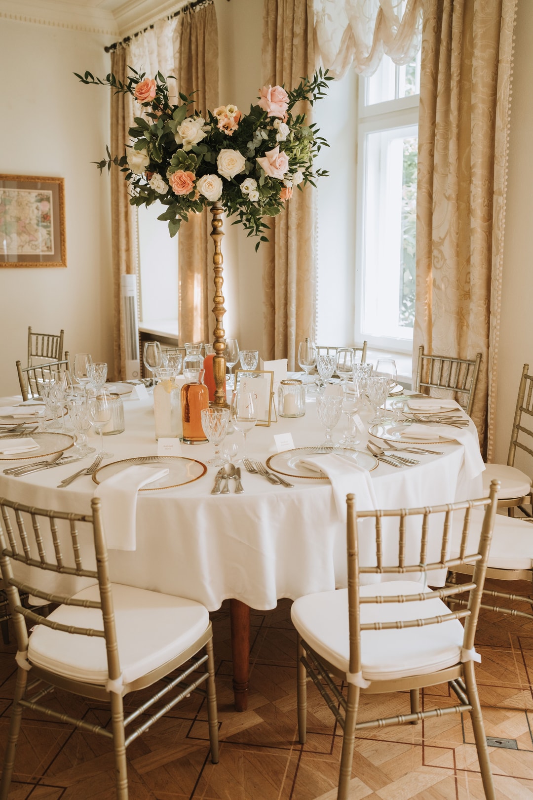 Aranżacja stołów weselnych z okrągłym stołem, białym obrusem, wysoka kompozycja kwiatowa na drewnianym postumencie z różami w jasnej palecie, kieliszkami i nakryciami stołu.