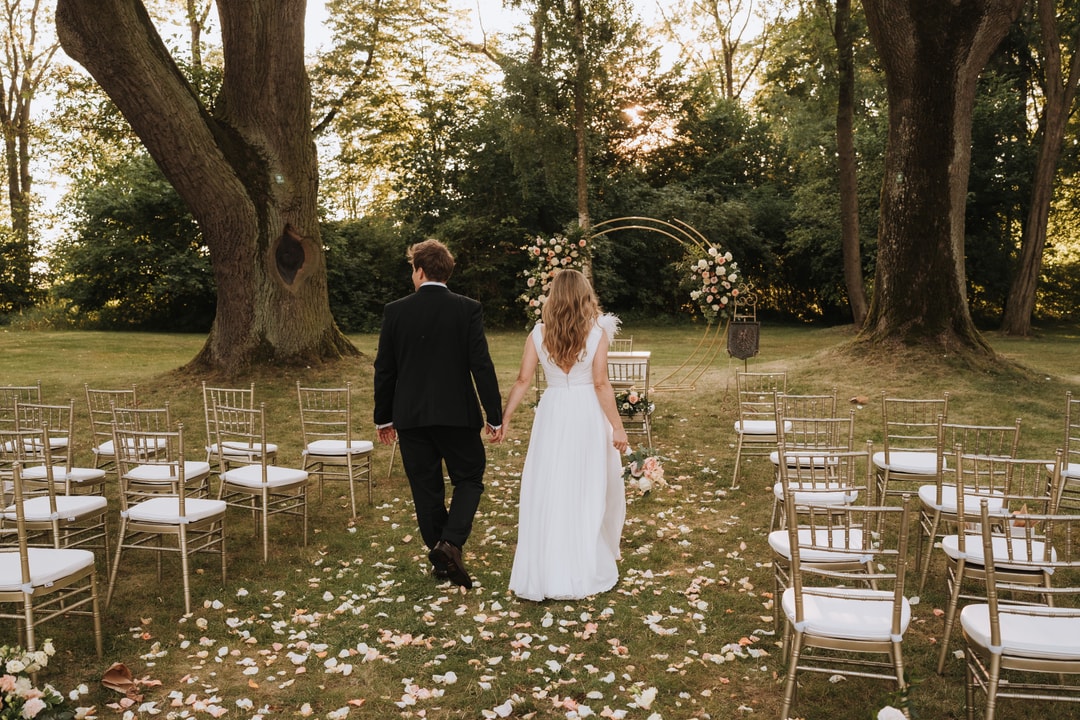 Para Młoda spacerująca aleję weselną między złotymi krzesłami, łuk kwiatowy z różowymi kwiatami w tle, naturalny krajobraz z dużymi drzewami.