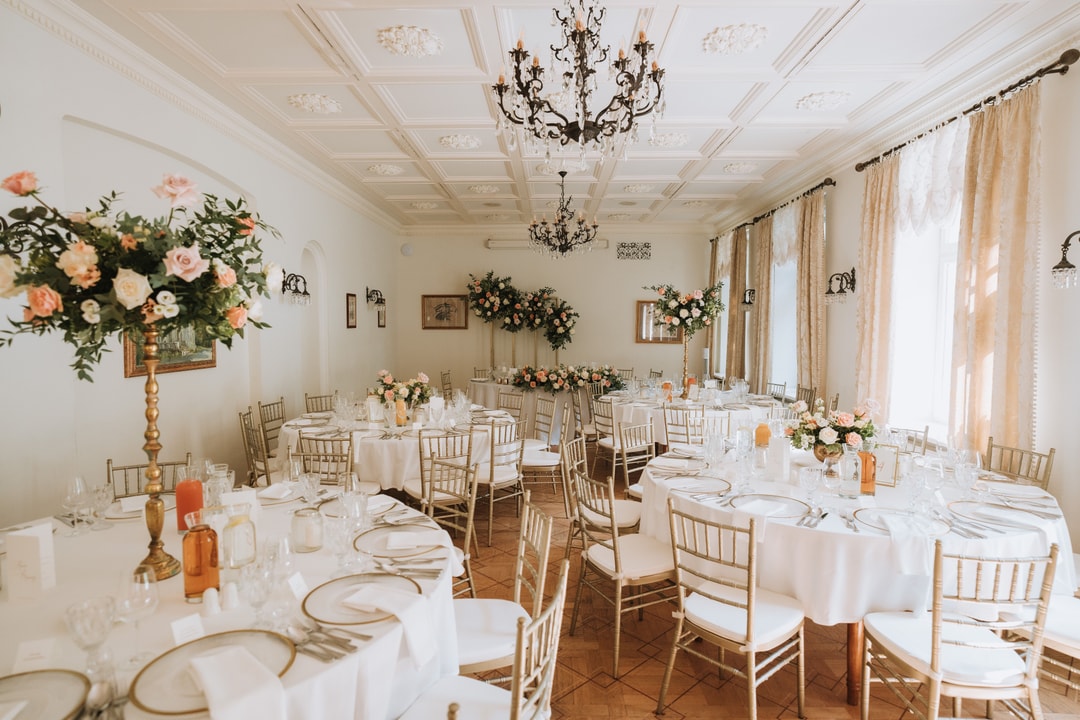 Elegancka sała weselna z dekoracjami sali weselnej - okrągłe stoły w białym obrusie, jasne kwiaty w композycях, złote chiavari, żyrandol, długie okna z beżowymi zasłonami.