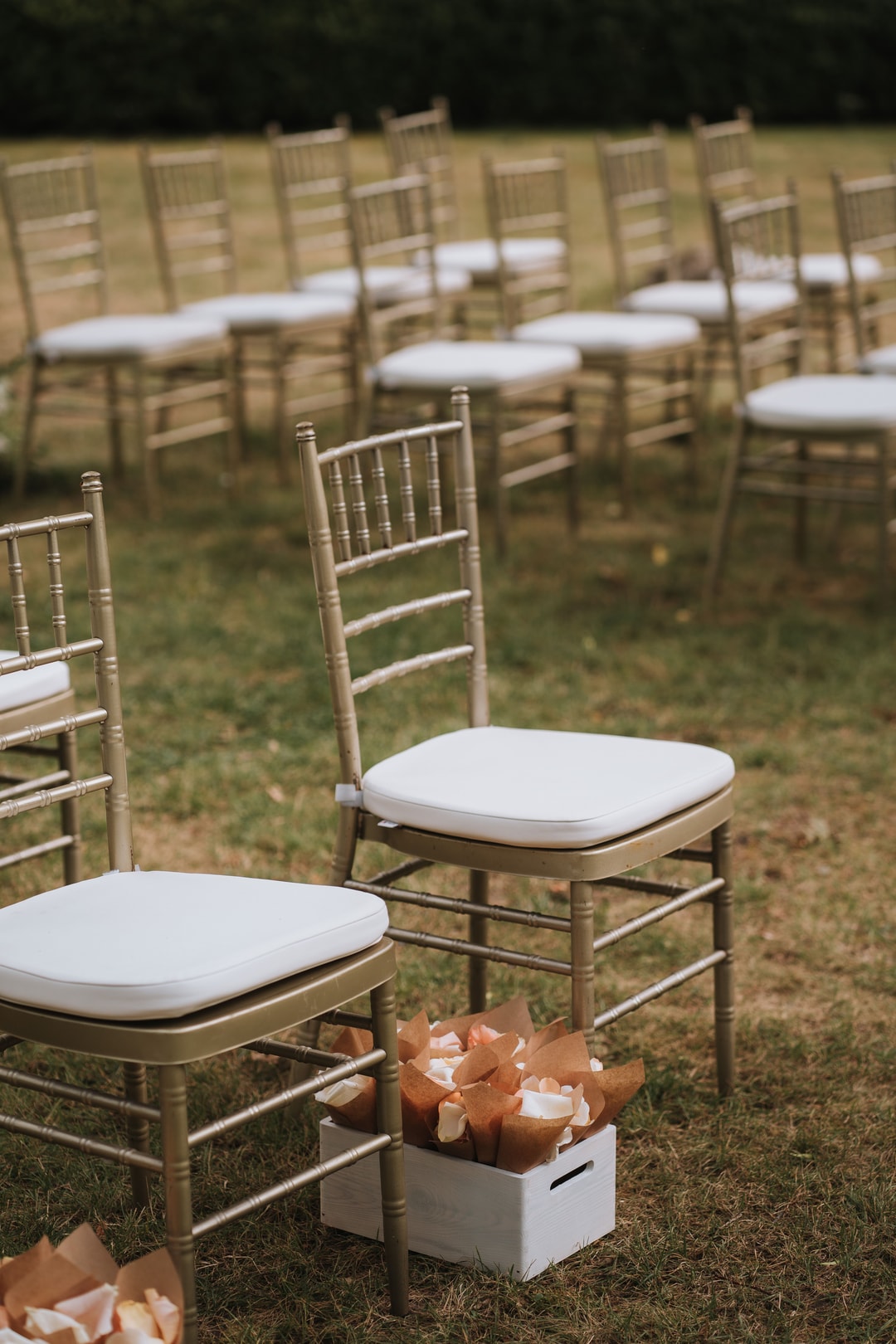 Rzędy krzeseł drewnianych z białymi poduszkami na trawniku, aranżacja stołów weselnych, kwiaty w pudełkach pomiędzy krzesłami