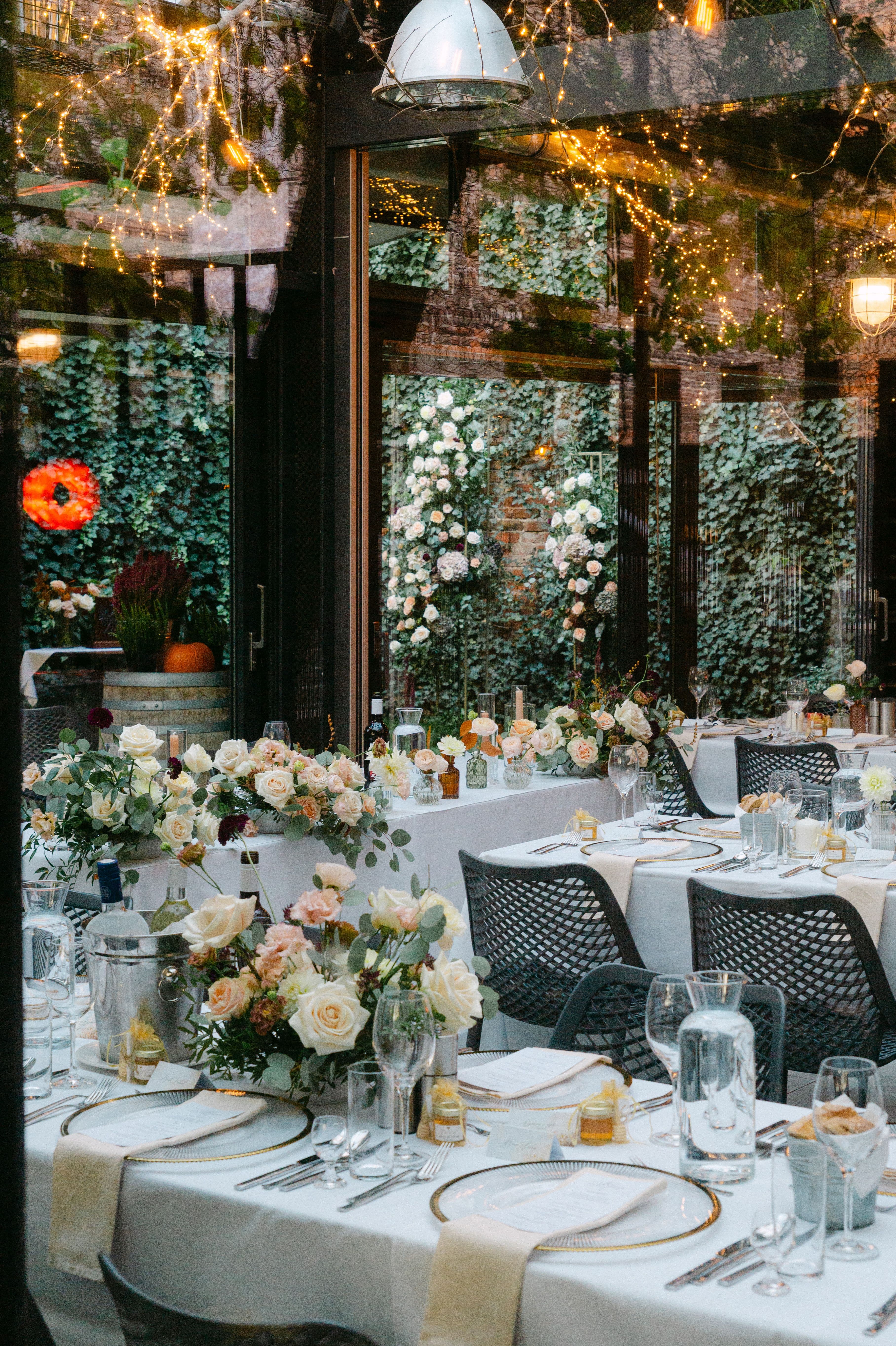 Aranżacja stołów weselnych z białymi i łososiowymi kwiatami w wazonach, białymi obrusami, czarnymi krzesłami i dekoracją ścianki kwiatowej z bluszczem.