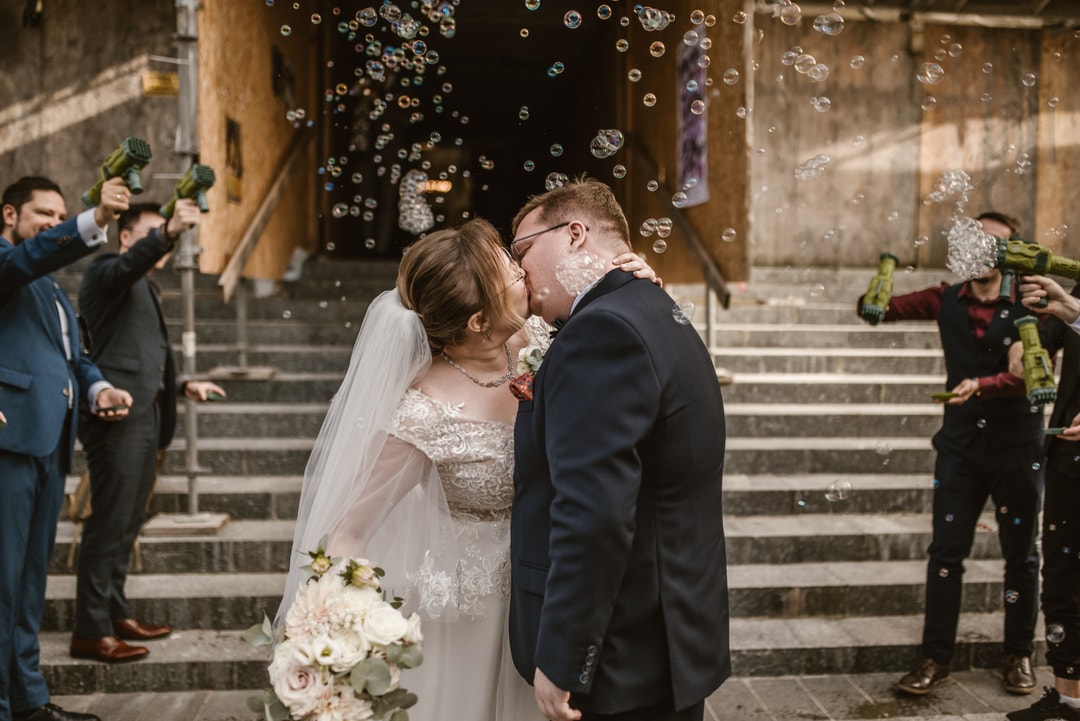 Para Młoda całuje się na schodach podczas cerementy ślubnej, otoczona bańkami mydlanymi i gośćmi z bukietami kwiatów.