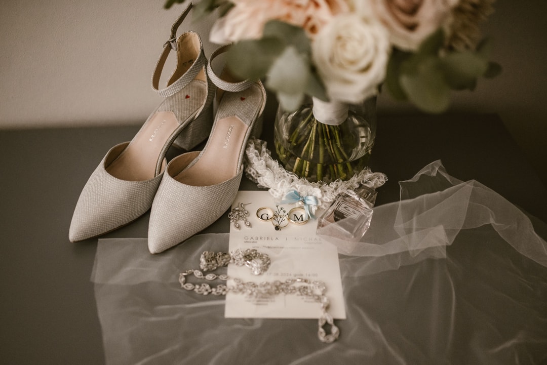 Buty ślubne w jasnej tonacji z metaliczną ozdobą, papeteria ślubna z monogramem oraz biżuteria i bukiet kwiatowy na tle