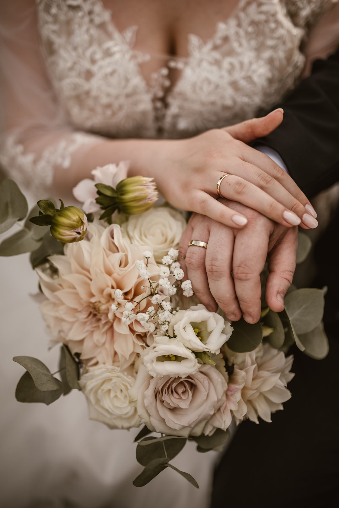 Ręce pary młodej z obrączkami ślubnimi na bukiecie z różami i kwiatami w jasnej palecie