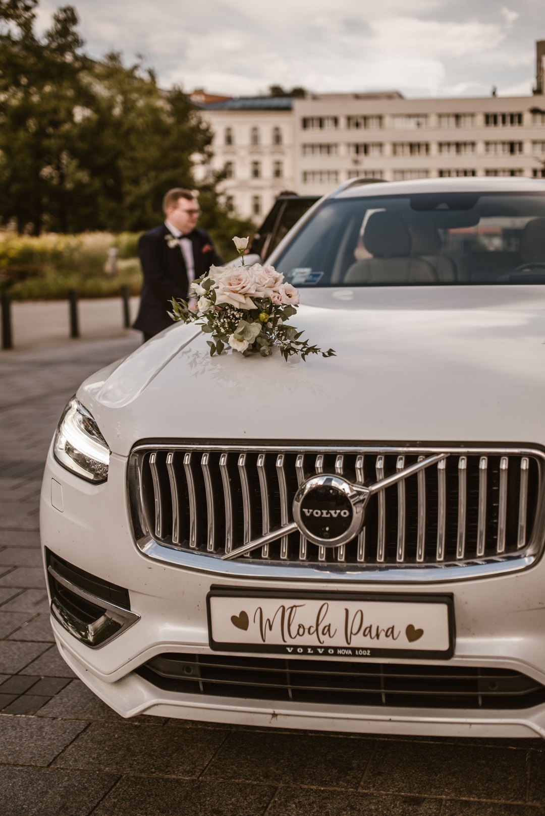 Biały samochód z композycją kwiatową na masce, w tle mężczyzna w garniturze, zabytkowa kamienica