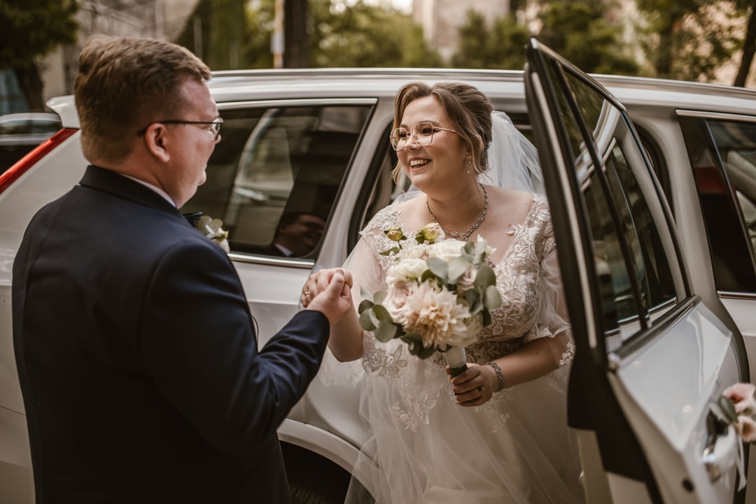 Panna młoda w białej sukni z bukietem kwiatów w jasnej palecie wychodzi z samochodu weselnego, uśmiechając się do pana młodego w czarnym garniturze.