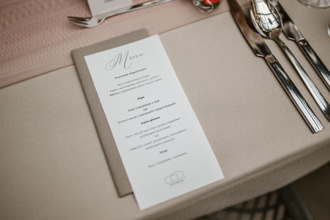 Papeteria ślubna - menu na stole weselnym z eleganckim pismem na białym tle, złożone na beżowym obrusie obok sztućców