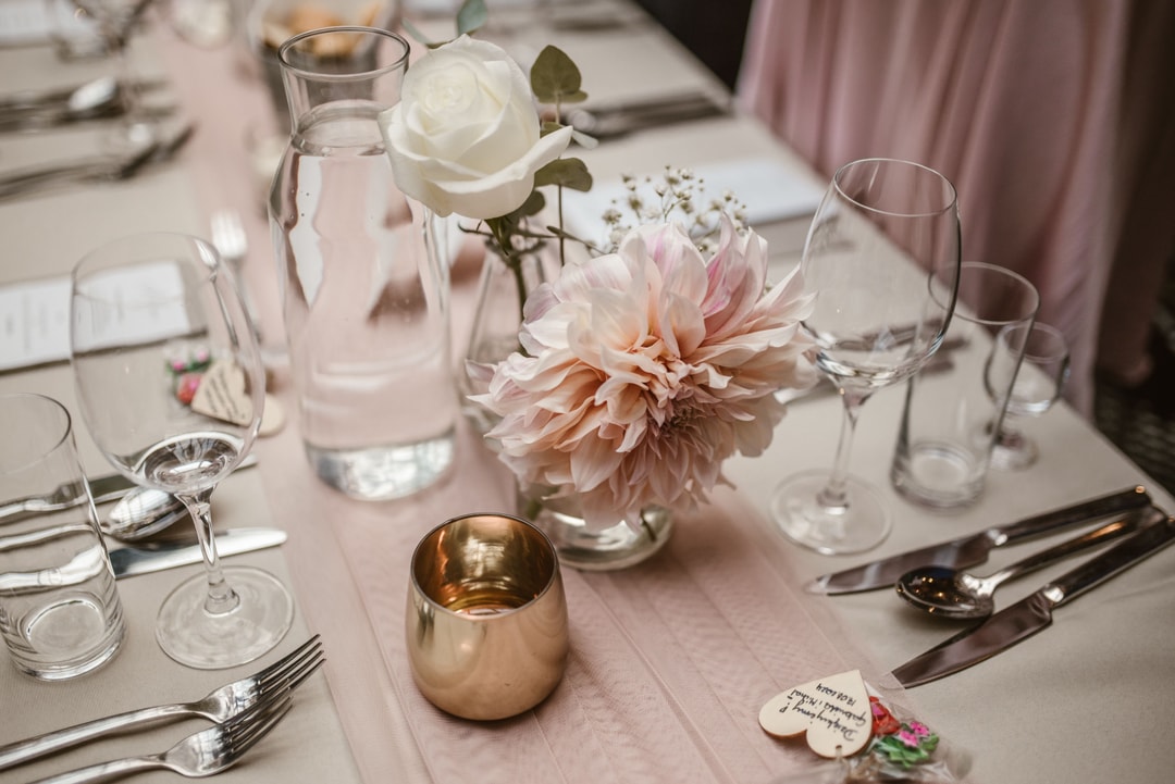 Dekoracja stołu weselnego z białą różą, pudrową dalią, szklankami i sztućcami na różowym obrusie