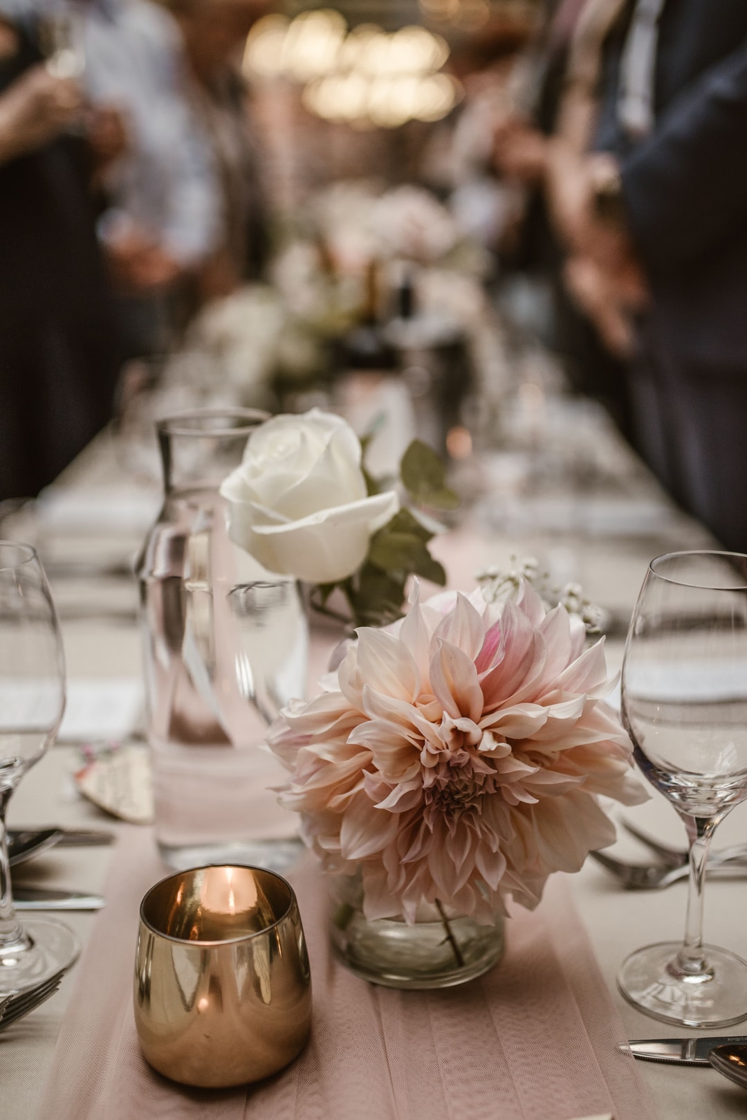 Zbliżenie na dekorację stołu z różową dalią, białą różą w szklanym wazoniku, złotym świecznikiem i kieliszkami, tło rozmyte z gośćmi