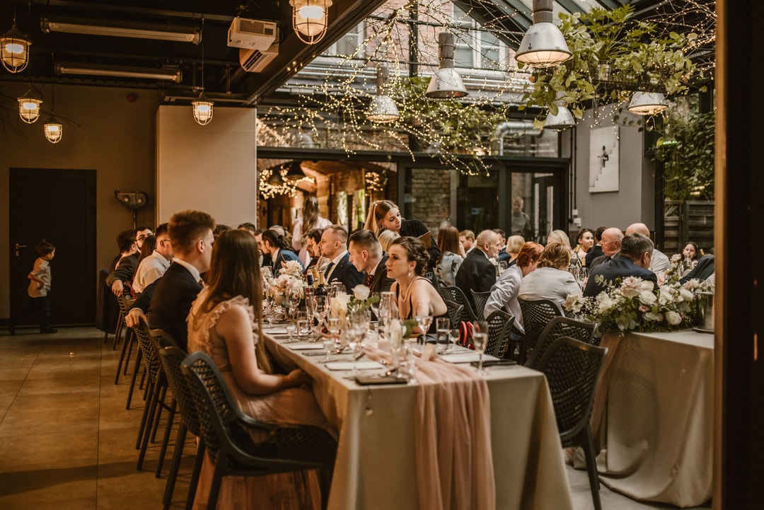 Aranżacja stołów weselnych z białymi obrusami, czarnymi krzesłami, kwiatowymi dekoracjami i zastawą. Goście siedzą wzdłuż długich stołów w eleganckiej sali z przemyślanym oświetleniem i zielenią.