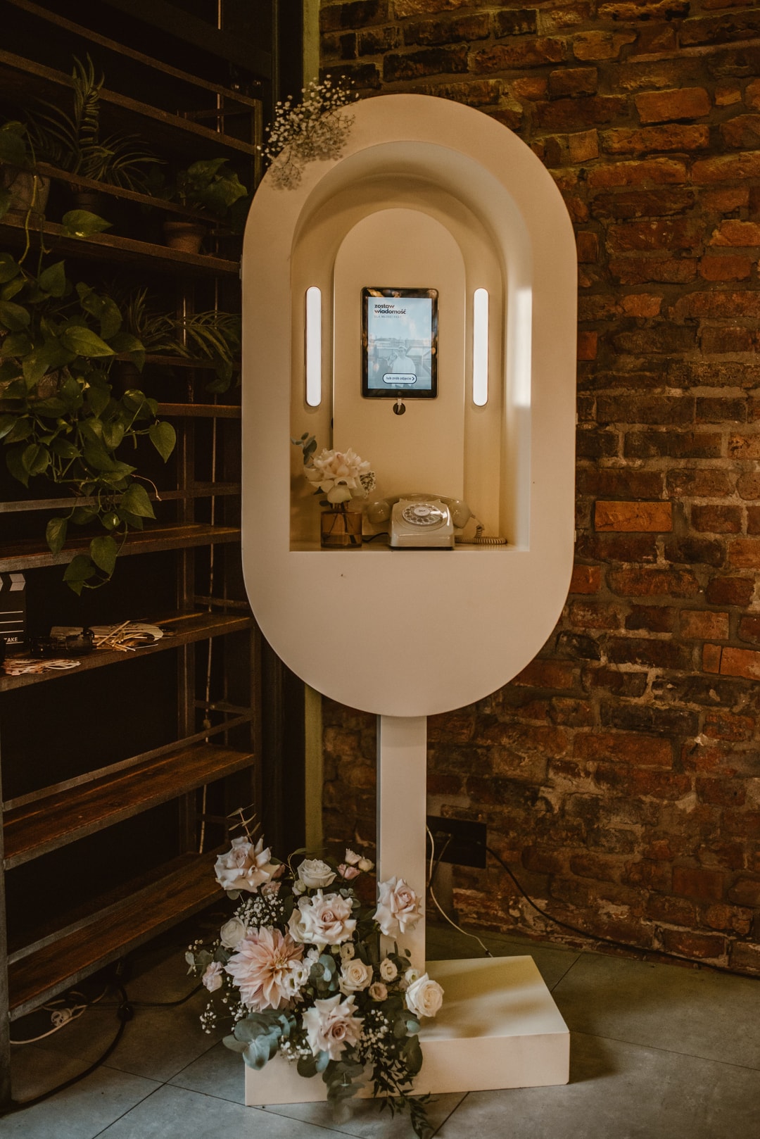 Nowoczesna dekoracja z białą obudową, kwiaty w jasnej palecie u podstawy, ekran, czerwona cegła w tle