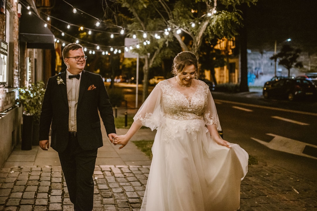 Para w strojach ślubnych spacerująca wieczorem po ulicy z girlandą świateł nad głowami.