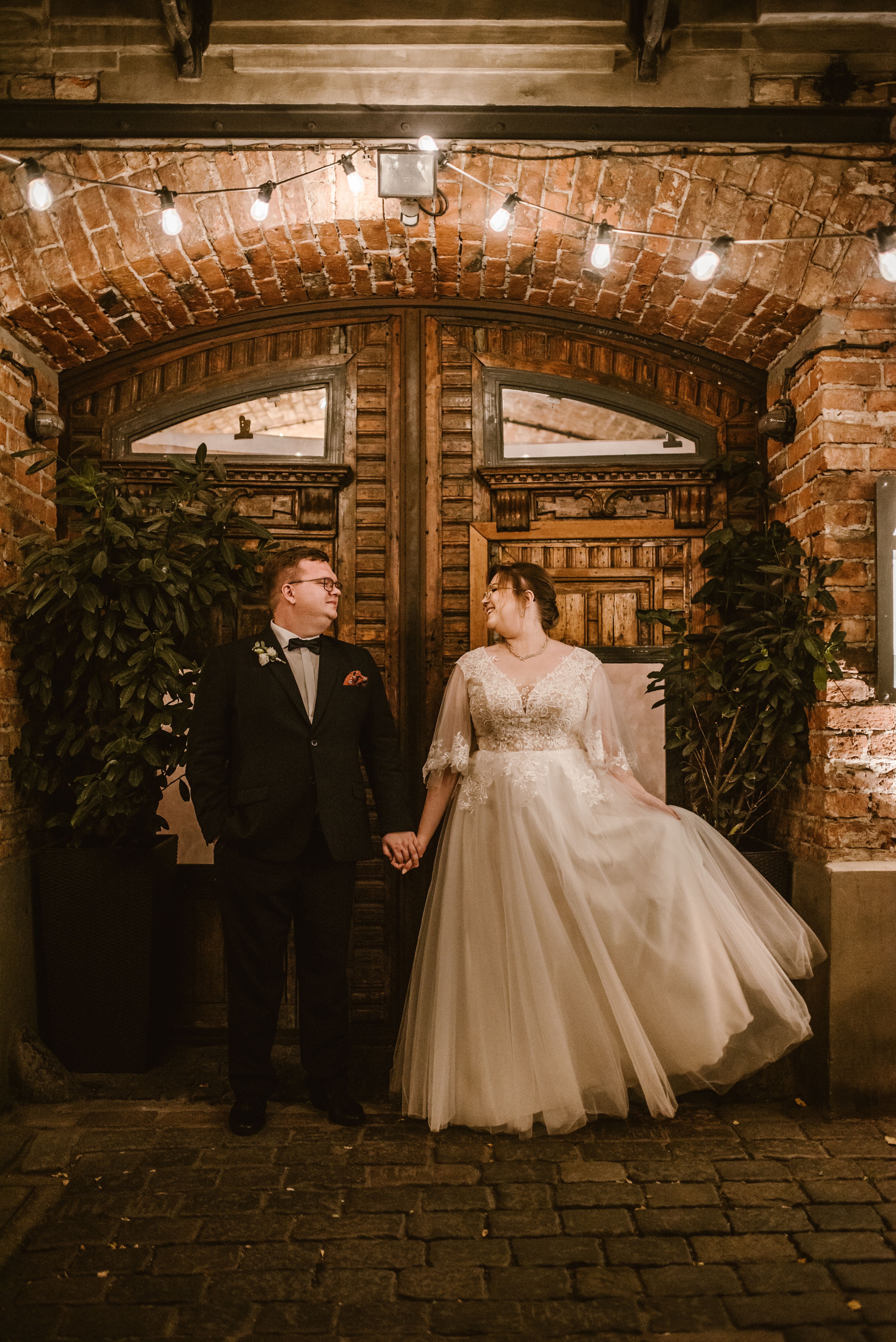 Para Młoda przy drewnianych drzwiach, mężczyzna w czarnym garniturze, kobieta w białej sukni ślubnej, zielone rośliny po bokach, żarówki nad wejściem.