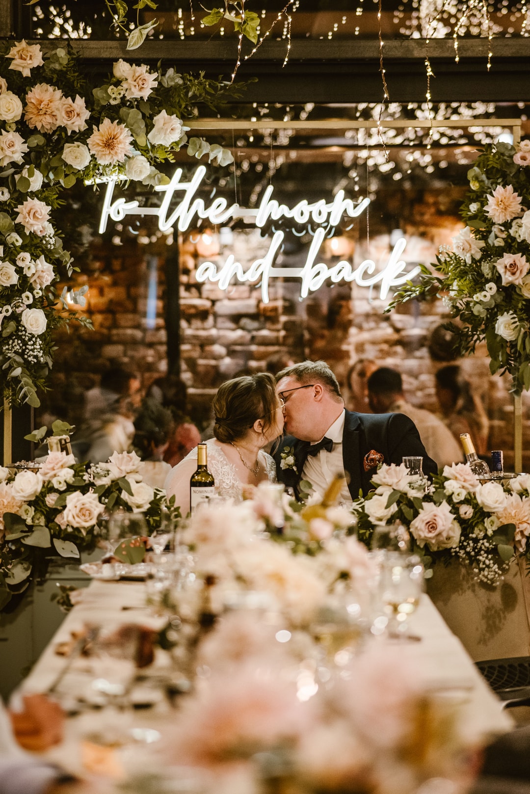 Para Młoda całuje się przy stole z dekoracją kwiatową, neon z napisem "to the moon and back" nad głowami, jasne kwiaty, gości w tle
