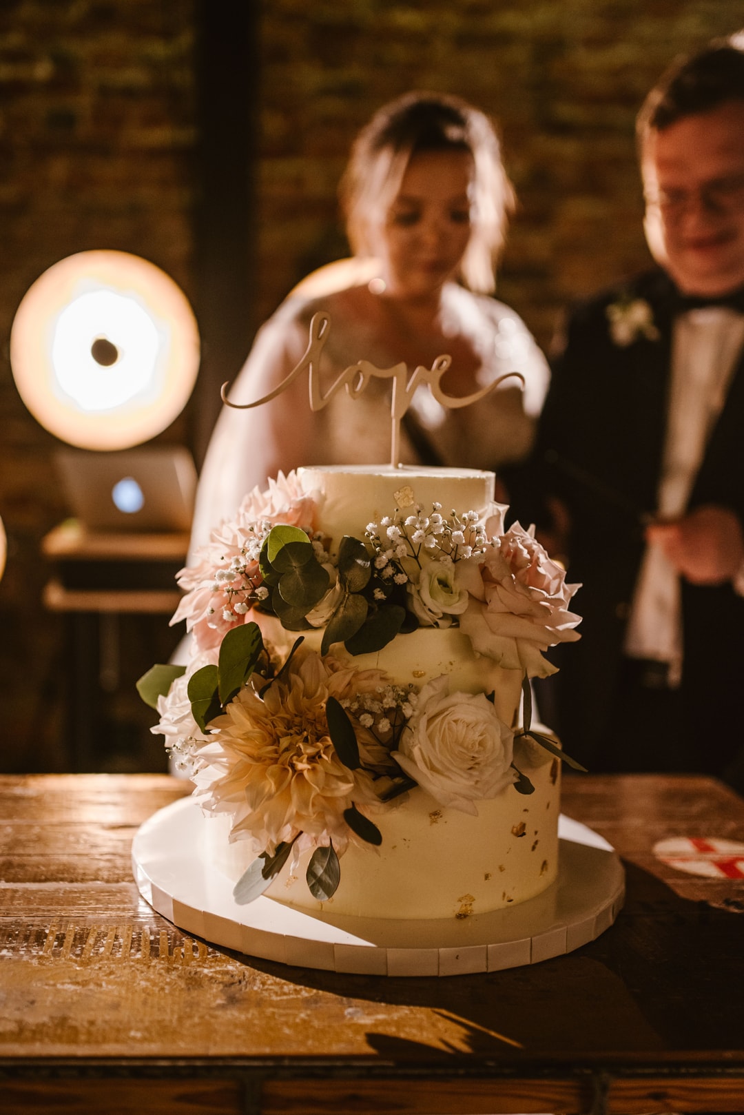 Dwupoziomowa kompozycja kwiatowa na stół z białym tortem, różowymi i kremowymi kwiatami, zielenią, napis love, w tle para gości w sali z ceglanym murem