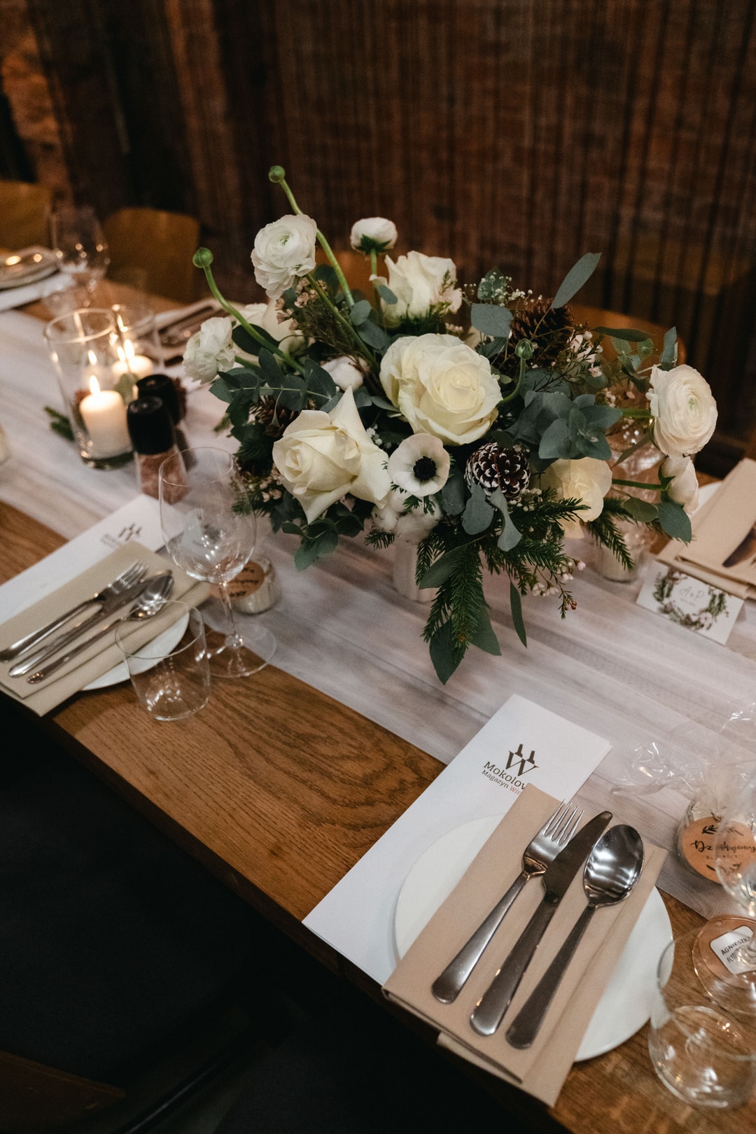 Dekoracja stołu z białymi różami, anemami, zielenią i oddychającymi kwiatami, srebrnym sztućcem, białymi talerzykami na drewnianym stole